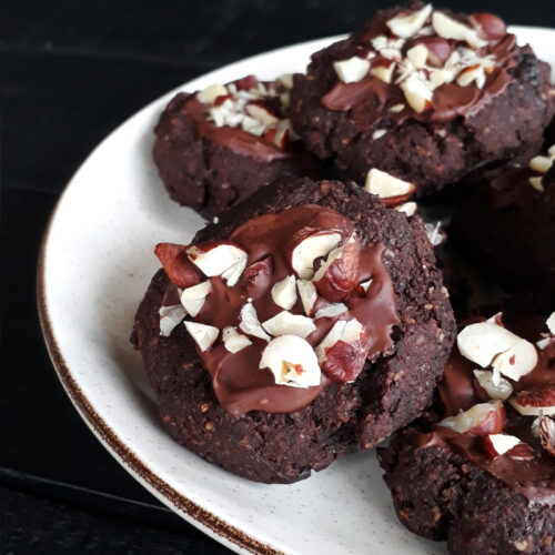 Vegan, gluten-free glazed hazelnut chocolate cookies garnished with hazelnut pieces, arranged on a plate.