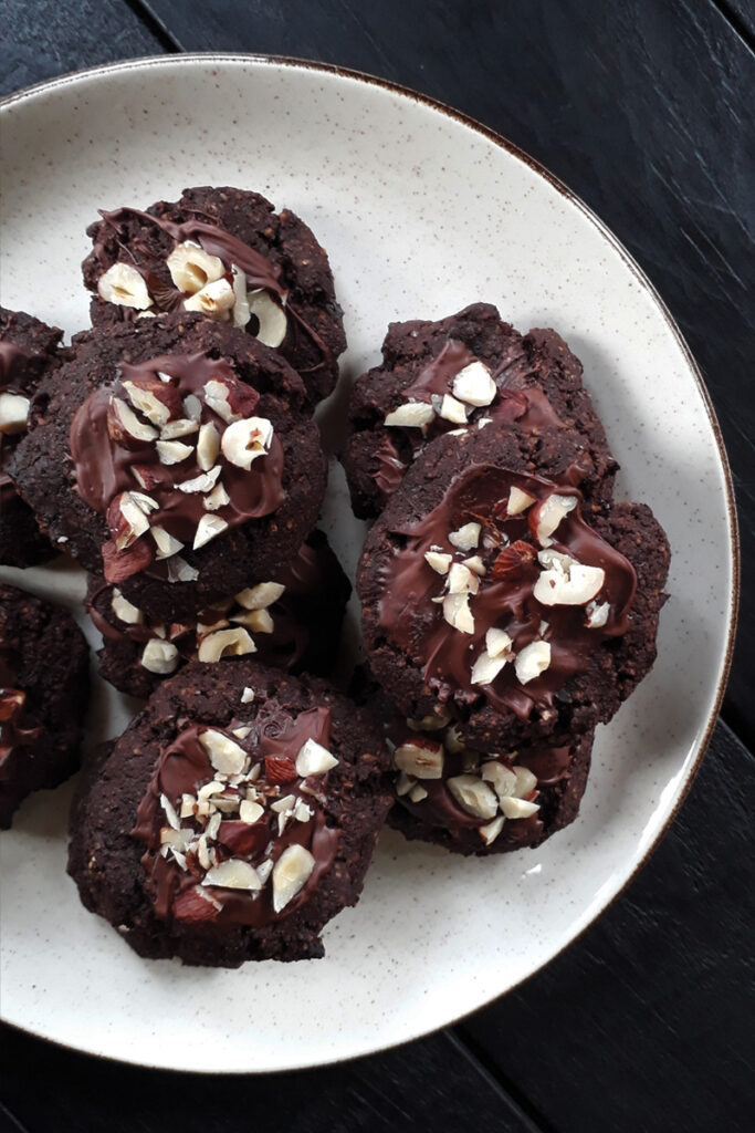Vegane Schoko Cookies (Glutenfrei, Ohne Getreide) 5 Glasierte vegane glutenfreie Haselnuss-Schoko-Cookies mit Haselnussstückchen, auf einem Teller angerichtet, aus Vogel-Perspektive fotografiert.