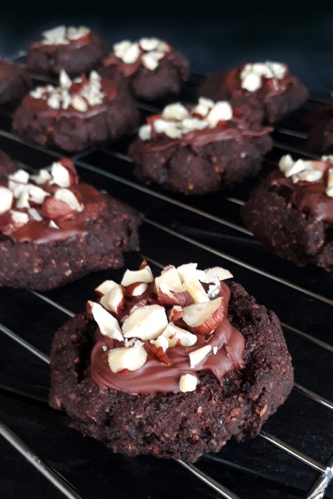 Vegane Schoko Cookies (Glutenfrei, Ohne Getreide) 4 Fertig glasierte Haselnuss-Schoko-Cookies, garniert mit kleinen Haselnussstückchen, auf einem Kuchengitter zum Abkühlen.